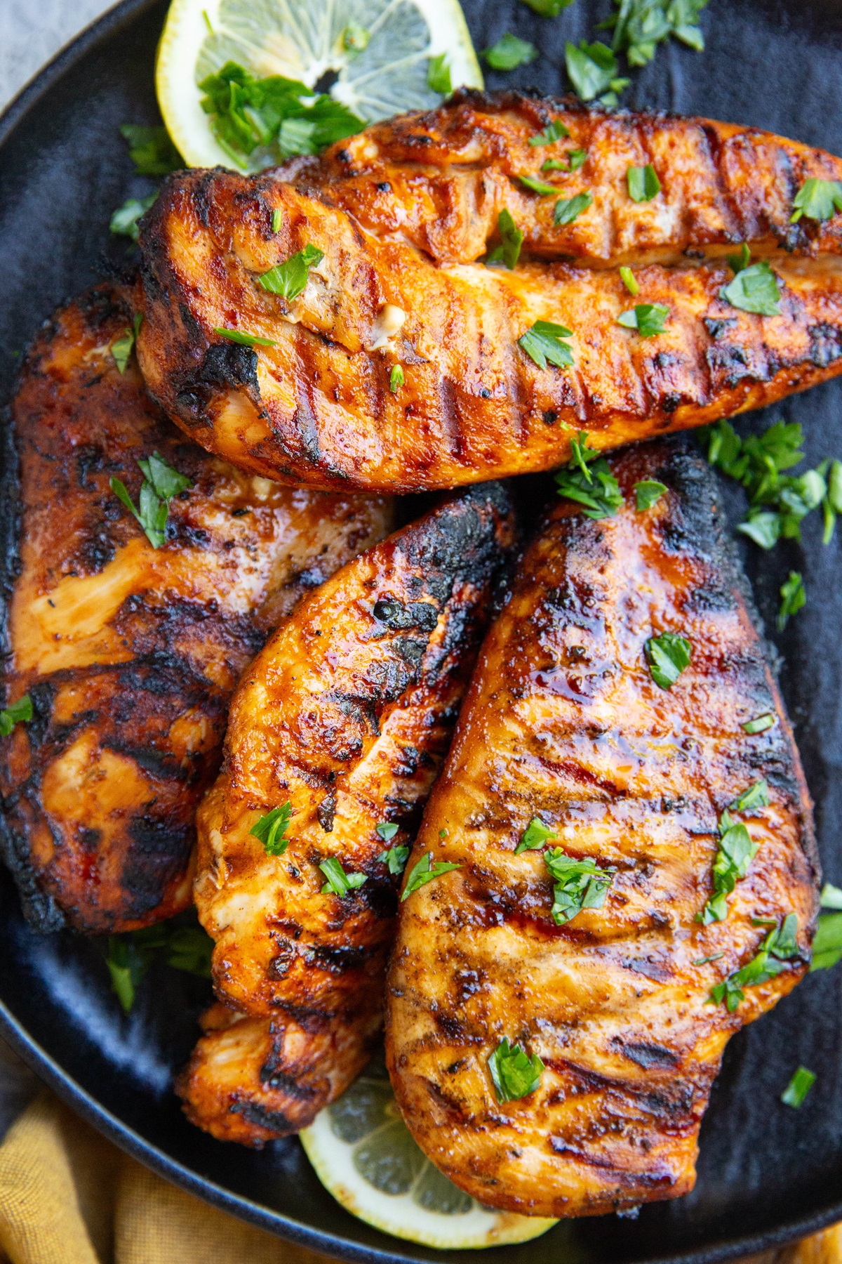 Black plate with four grilled chicken breasts sprinkled with parsley and slices of lemon to the side.
