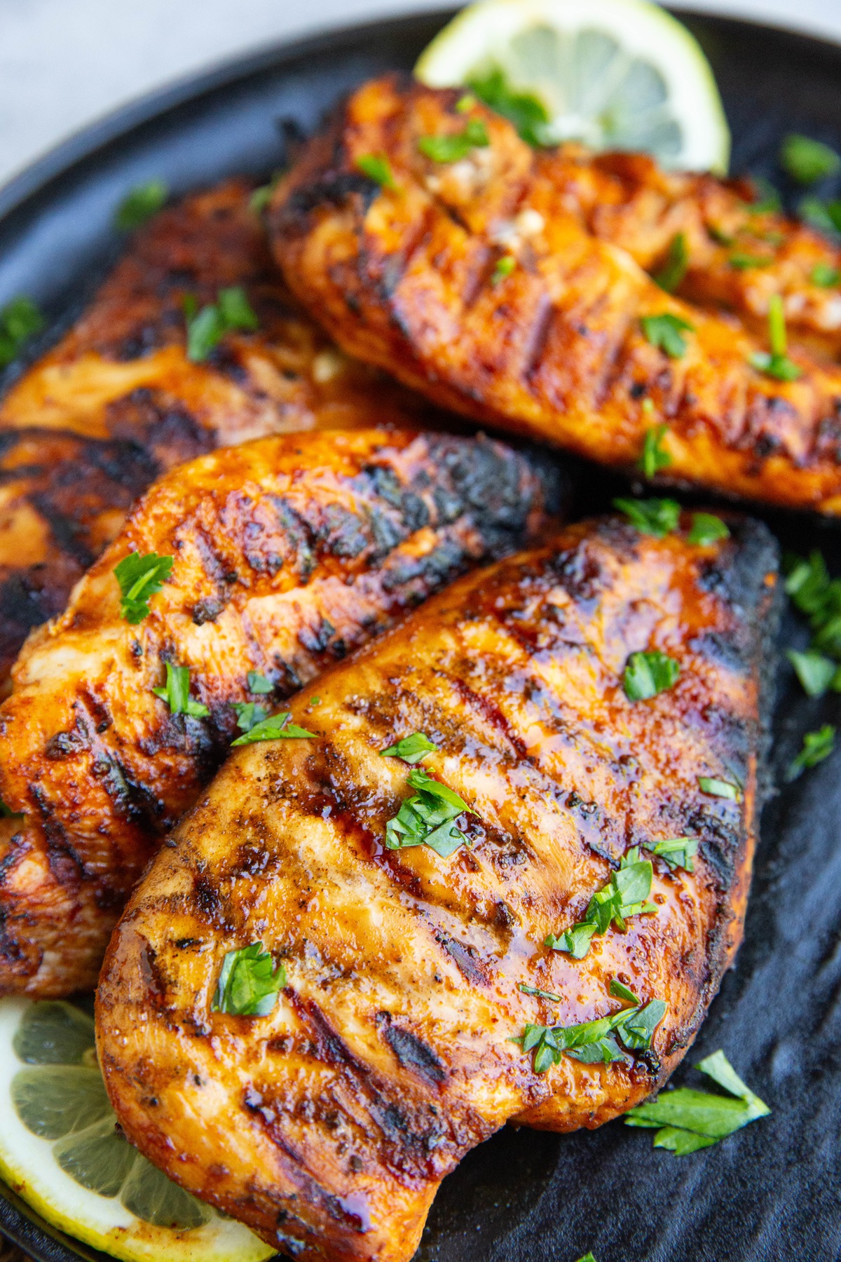 Four grilled chicken breasts on a black plate, sprinkled with fresh parsley and sliced lemons to the side.