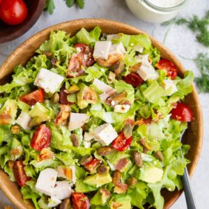 Chicken bacon avocado ranch salad in a wooden bowl with ranch dressing to the side and a bowl of tomatoes to the side.