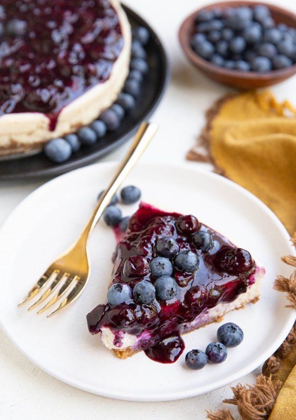 Slice of blueberry cheesecake on a white plate with the rest of the cheesecake in the background.