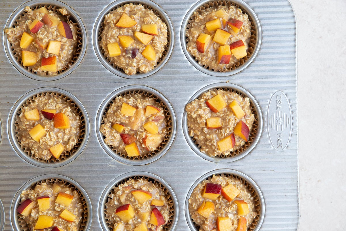 Peach oatmeal muffin mixture in a muffin tray with chopped peaches on top. Ready to go into the oven.