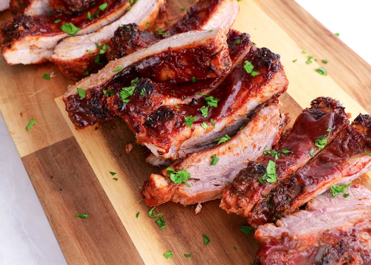 Cutting board with rack of ribs cut into slices, sprinkled with fresh parsley.