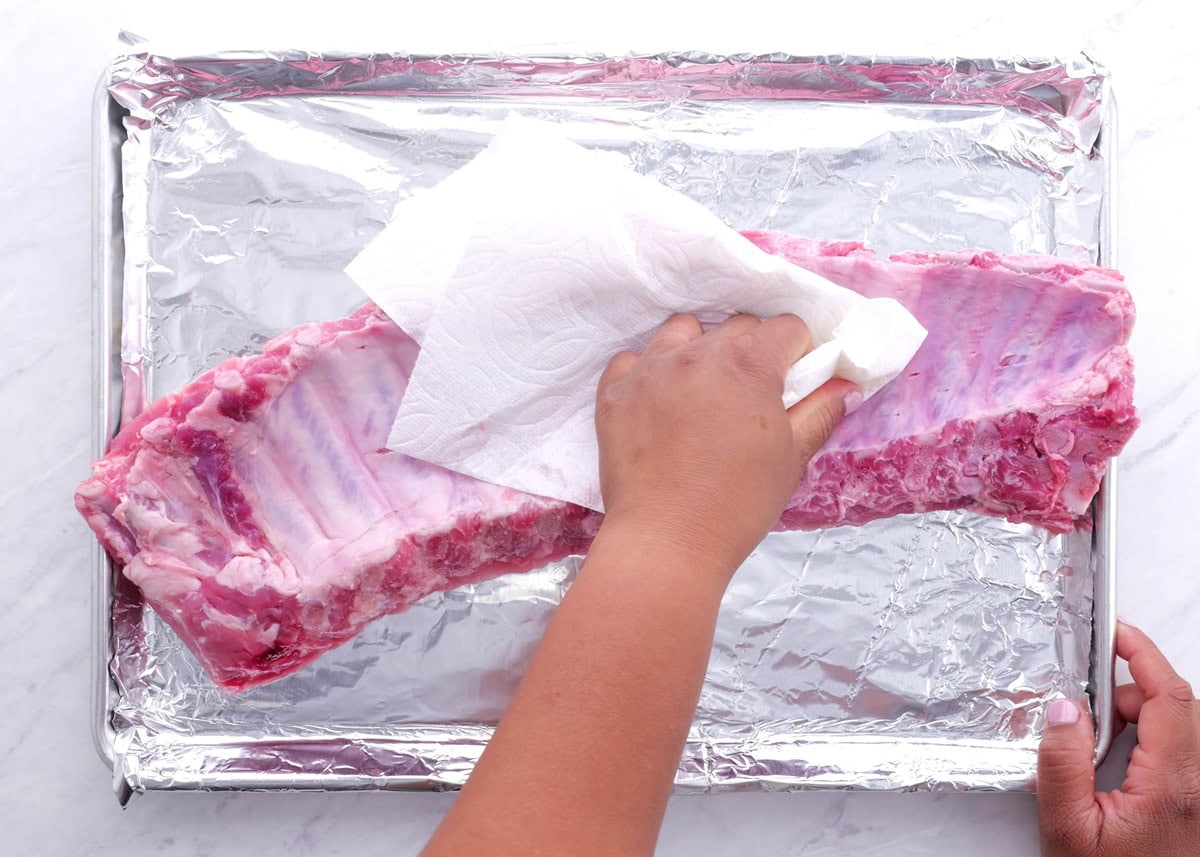 Hand patting a rack of ribs with a paper towel to remove moisture.