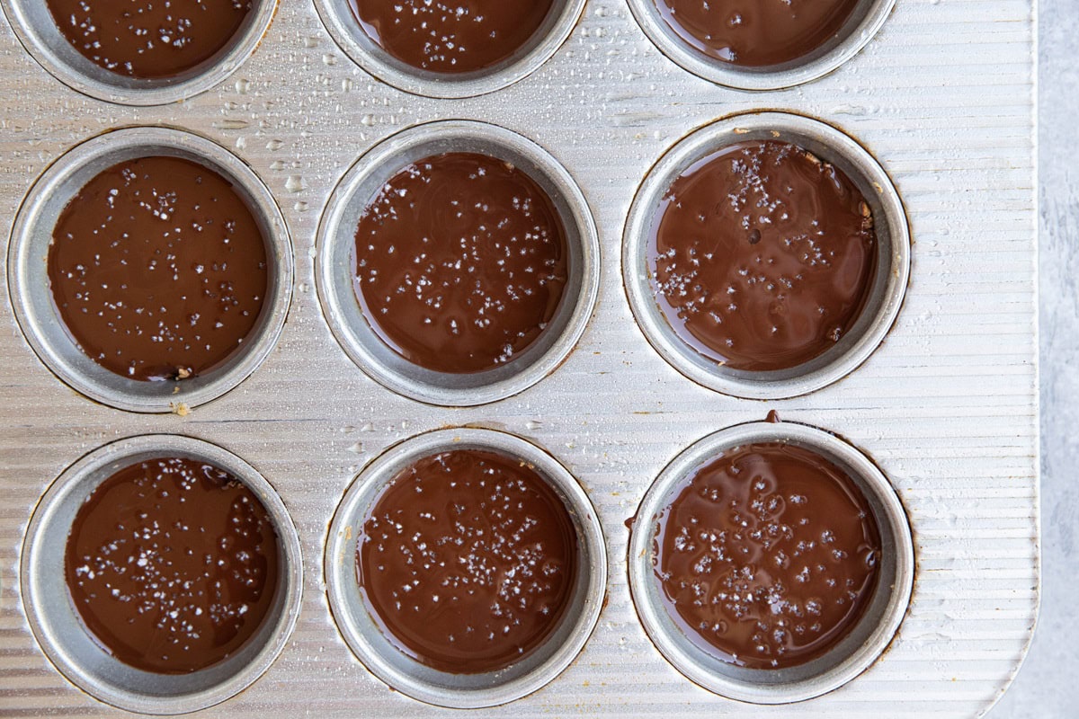 Muffin tin with melted chocolate on top of almond butter layer to make almond butter cups.