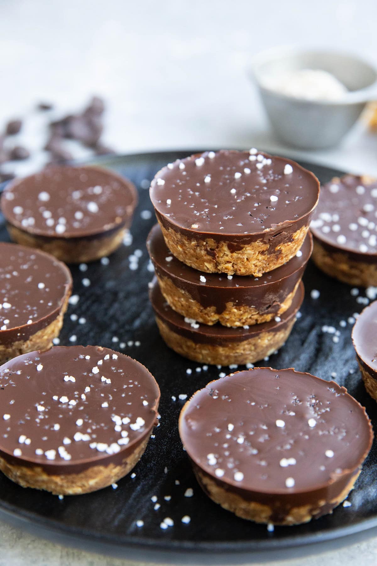 Stack of almond butter cups on a black plate with more cups all around.