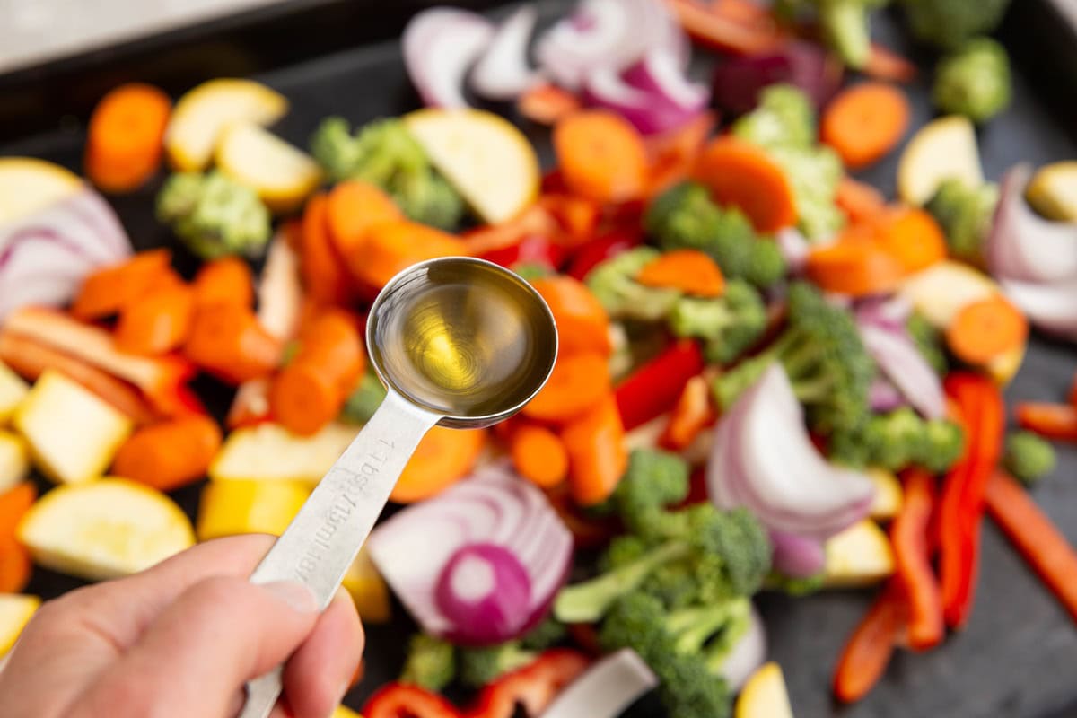 Hand holding a tablespoon of avocado oil over a sheet pan of vegetables.