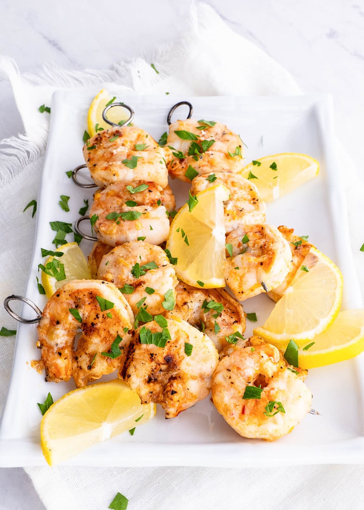 Grilled shrimp skewers on a white platter sprinkled with fresh parsley with lemon slices all around.