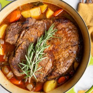 Pot roast in a Dutch oven with fresh herbs and vegetables.