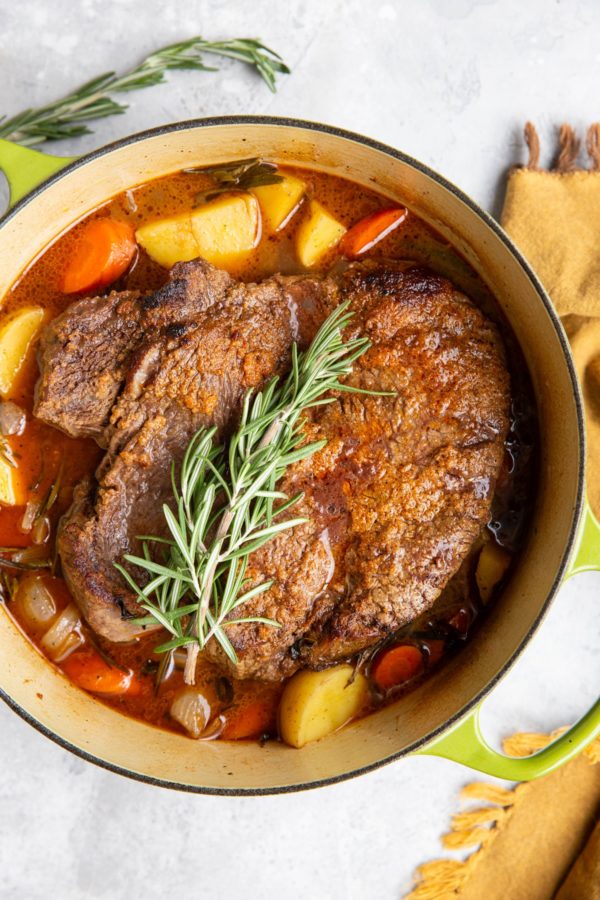 Pot roast in a Dutch oven with fresh herbs and vegetables.