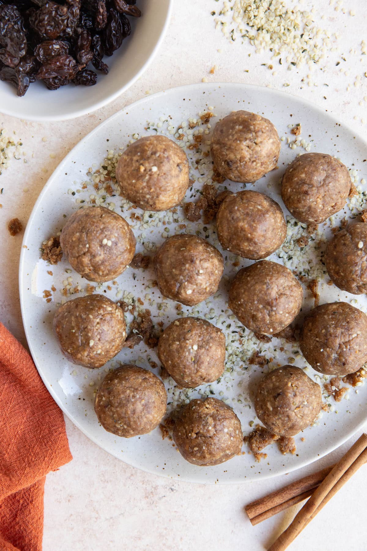 White plate of cinnamon raisin energy balls with cinnamon sticks, hemp seeds and raisins around.