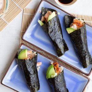 Four salmon hand rolls on plates with sauces to the side, ready to eat.