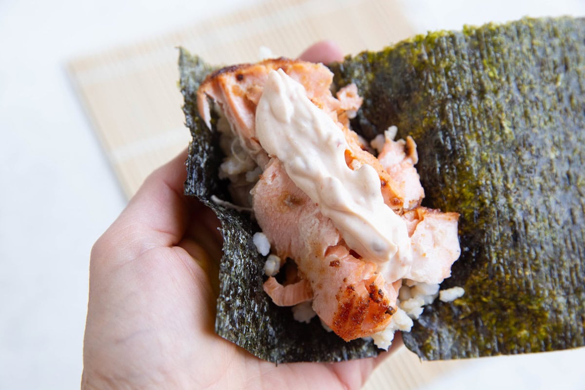 Hand holding seaweed wrapper with rice, cooked salmon and sauce.