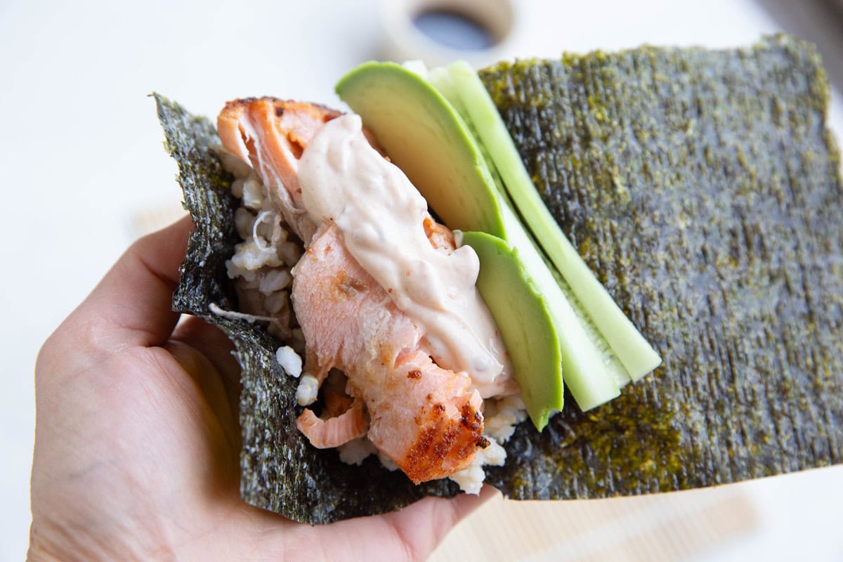 Hand holding a seaweed sheet with rice, salmon, sauce, avocado, and cucumber.