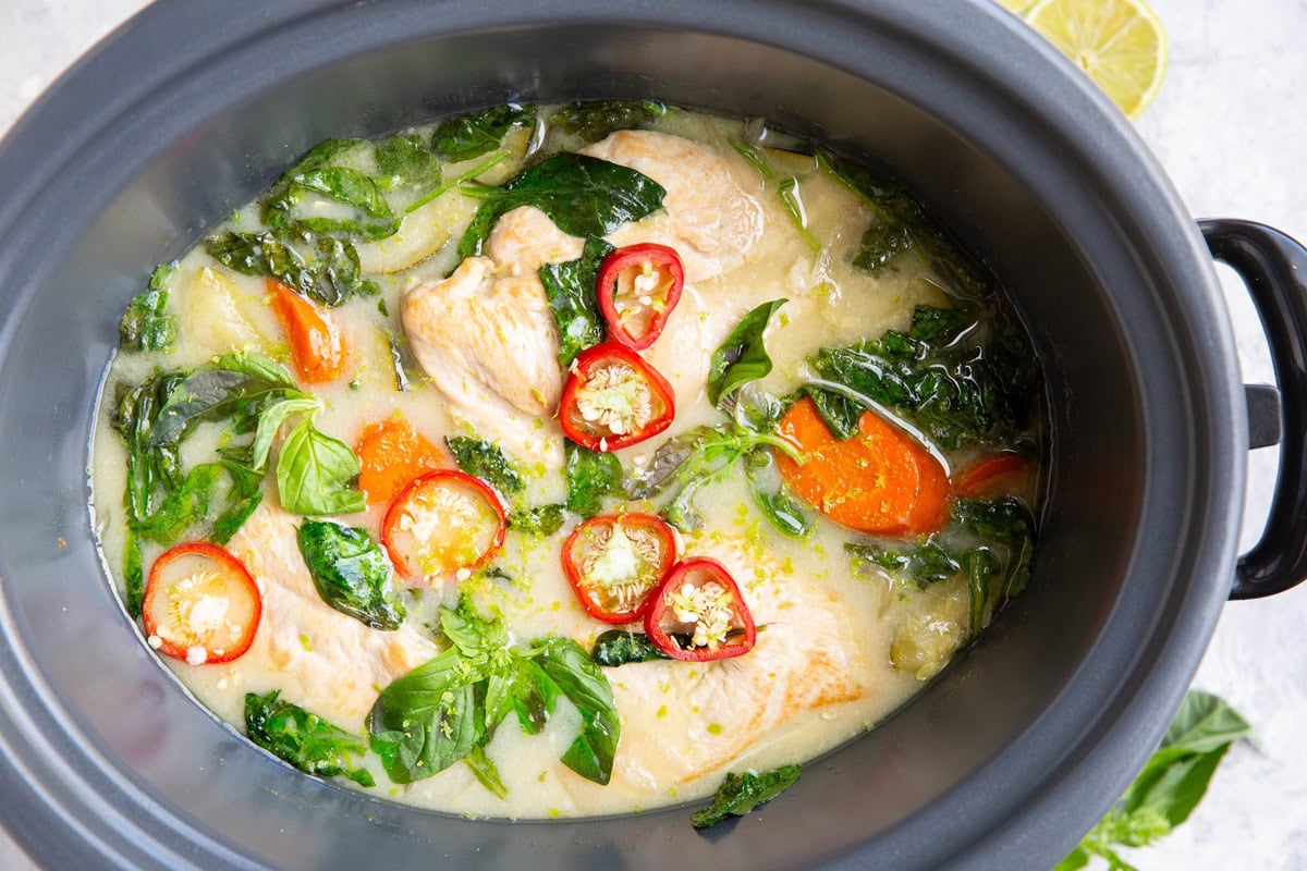 A slow cooker filled with chicken and creamy sauce with red chilis and basil on top and fresh lime to the side.