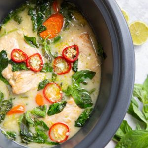 Crock pot full of chicken and sauce with basil and red chilies on top.