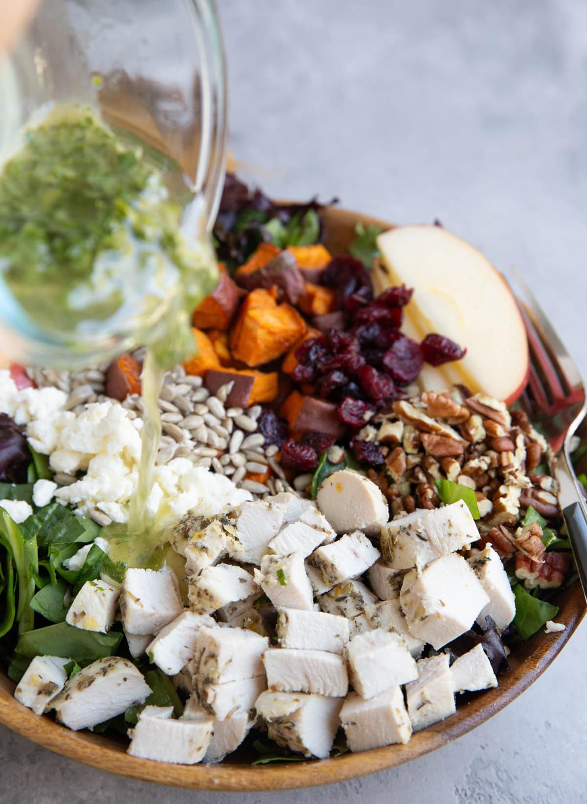 Wooden bowl of chicken sweet potato salad with a hand pouring dressing into the bowl.