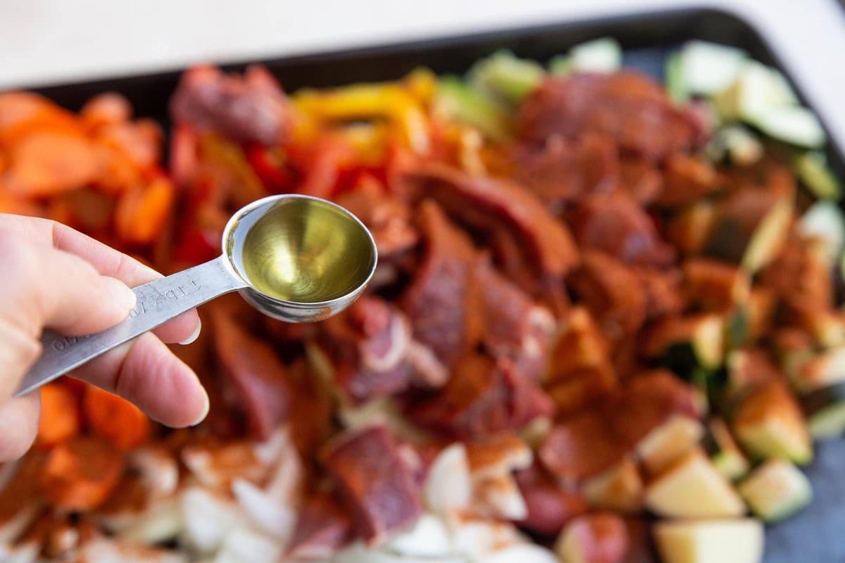 Baking sheet full of steak and vegetables with a hand holding a tablespoon of avocado oil.