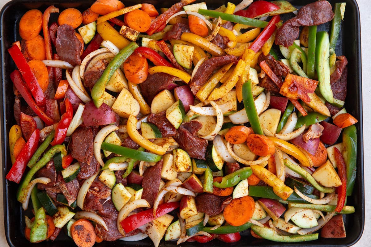 Raw beef and vegetables seasoned with chili powder and oil on a large baking sheet, ready to go into the oven.