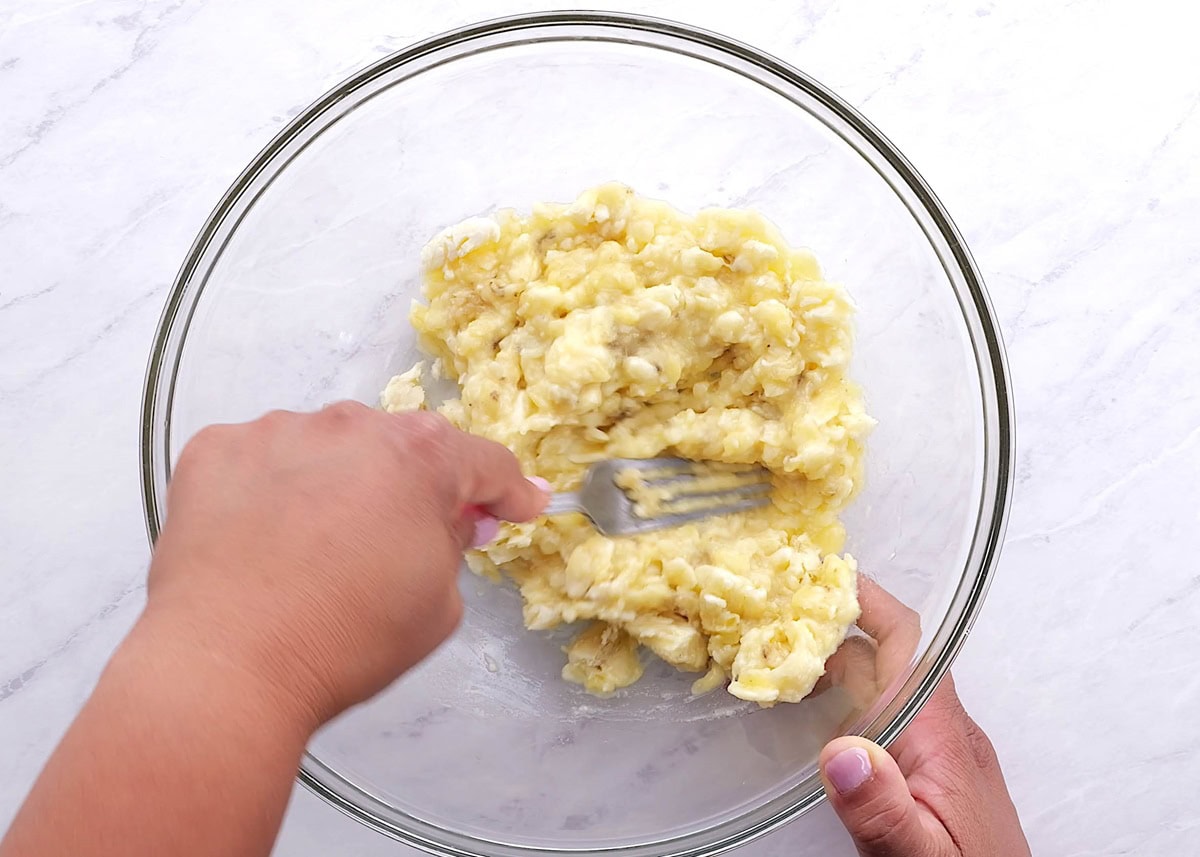 Hands mashing ripe bananas in a mixing bowl.