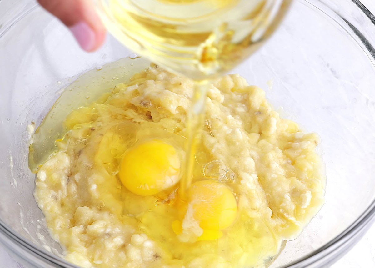 Pouring oil into a mixing bowl with mashed bananas and eggs.