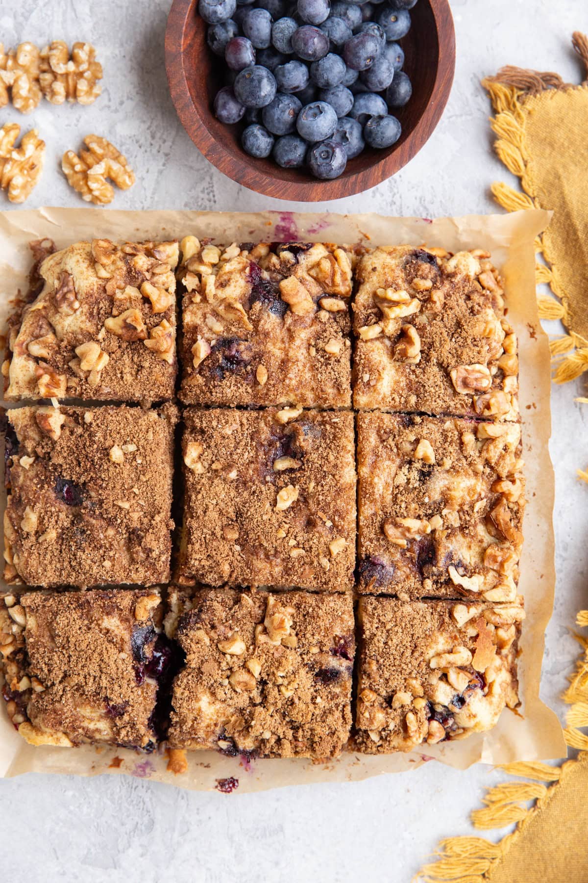 Yogurt coffee cake on a sheet of parchment paper, cut into individual slices with a bowl of fresh blueberries and walnuts all around.