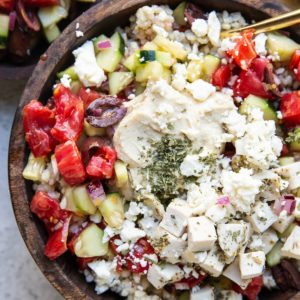 Wooden bowl full of chicken, rice, hummus, and cucumber tomato salad with a gold spoon and a bowl of salad to the side.