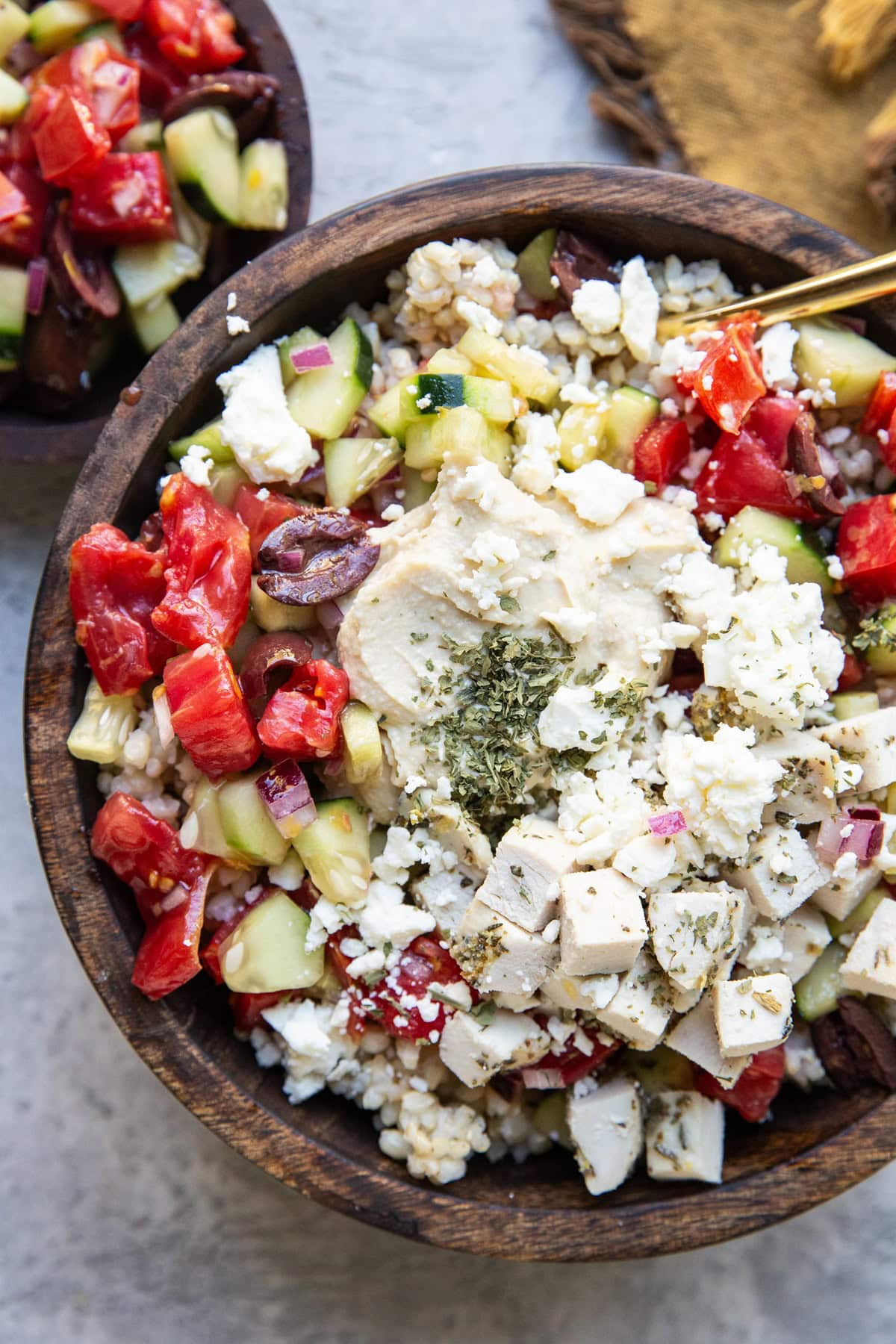 Wooden bowl full of chicken, rice, hummus, and cucumber tomato salad with a gold spoon and a bowl of salad to the side.