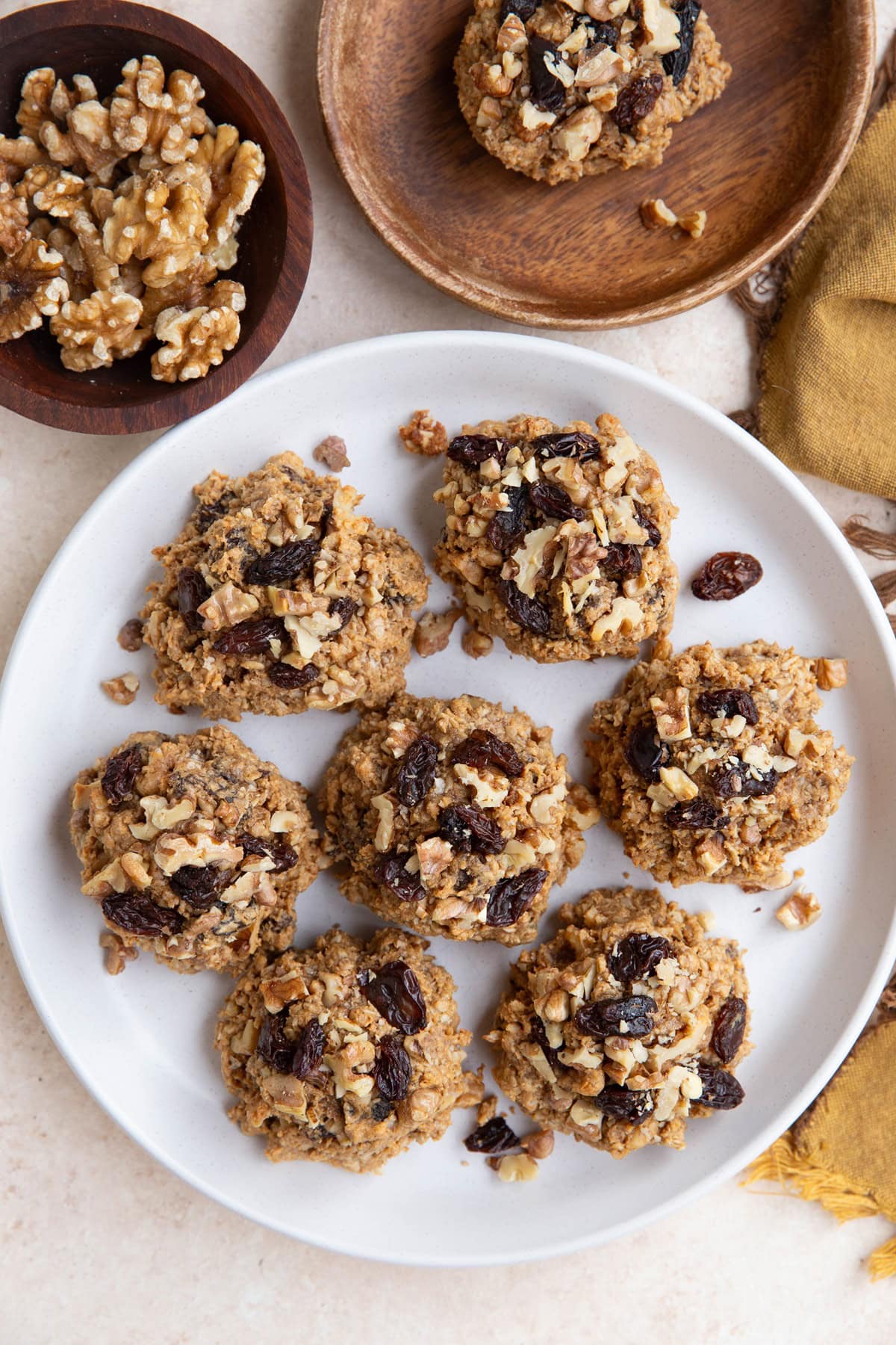 White plate of oatmeal raisin cookies with a wooden plate to the side and a bowl of walnuts.