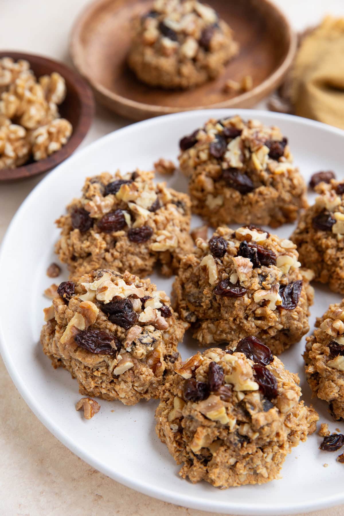 White plate of oatmeal raisin cookies with another cookie in the background and a bowl of walnuts.