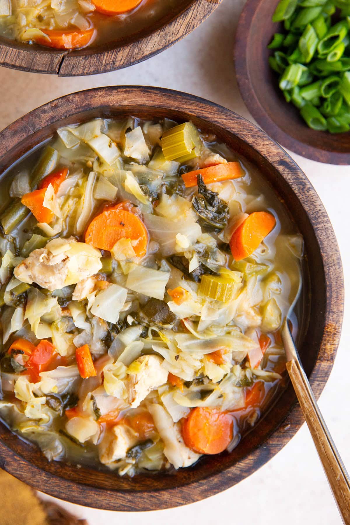 Two wooden bowls full of chicken vegetable soup with a bowl of green onions to the side, ready to eat.