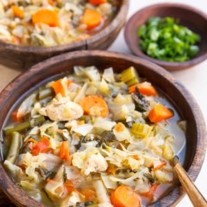 Chicken and cabbage soup in two wooden bowls with a bowl of green onions to the side, ready to eat.