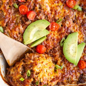 Large white casserole dish full of cheesy rice and beef casserole with a wooden spoon taking a scoop out to serve.