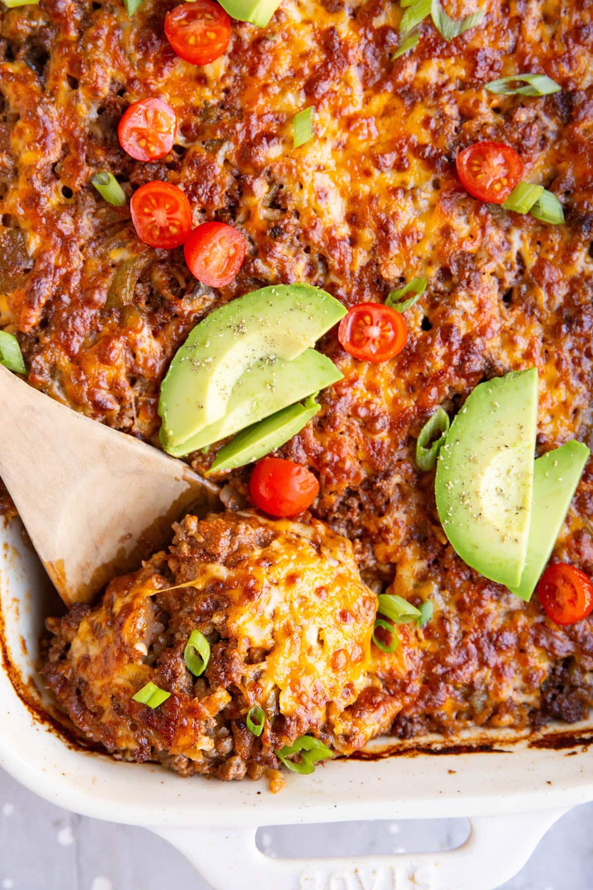 Large white casserole dish full of cheesy rice and beef casserole with a wooden spoon taking a scoop out to serve.