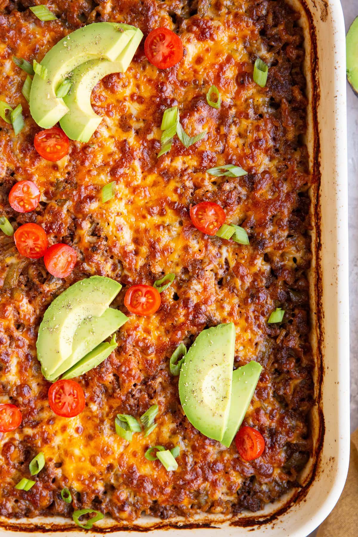 White baking dish of ground beef and rice enchilada casserole with sliced avocado and tomatoes on top.