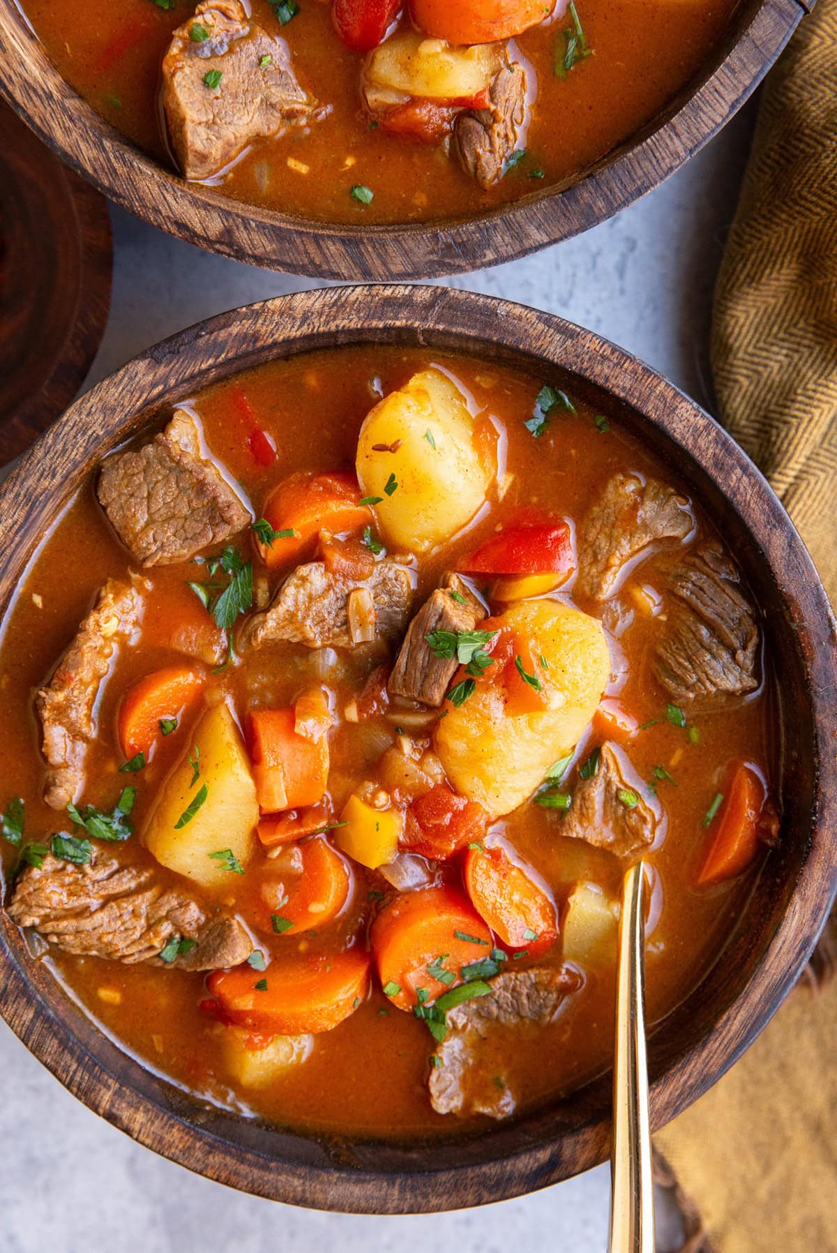 Two wooden bowls full of Hungarian beef goulash with a gold spoon and napkin to the side