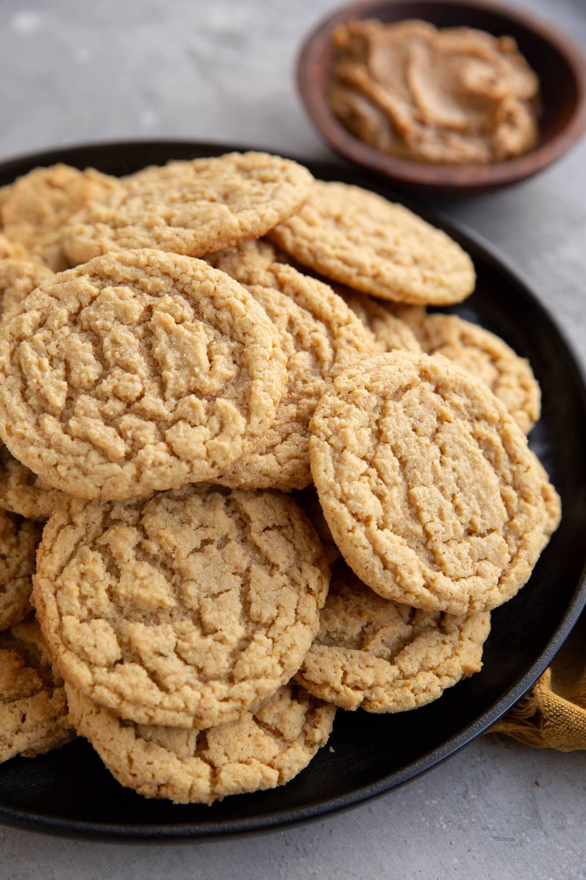 Big stack of peanut butter cookies on a plate with a bowl of peanut butter in the background, ready to eat.