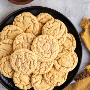 Black plate with a stack of almond flour peanut butter cookies on top with a wooden bowl of peanut butter to the side.