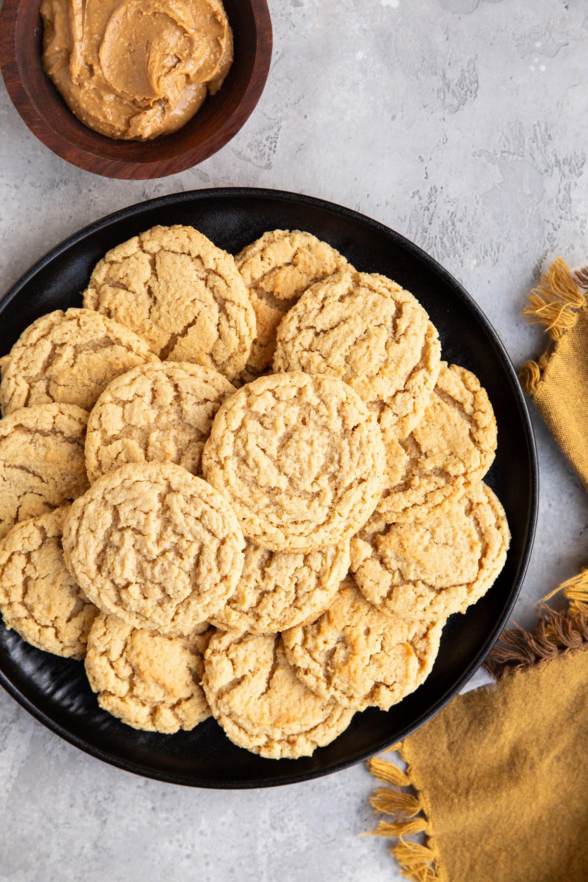 Black plate with a stack of almond flour peanut butter cookies on top with a wooden bowl of peanut butter to the side.