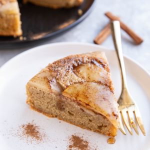 Slice of pear cake on a white plate with gold fork and cake in the background.