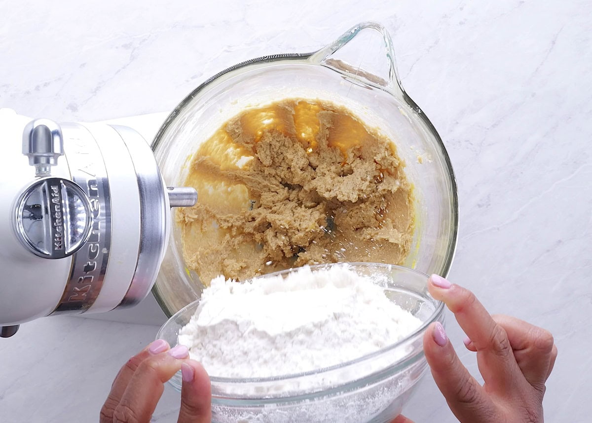 Hands pouring flour mixture into a stand mixer to make cookie dough.