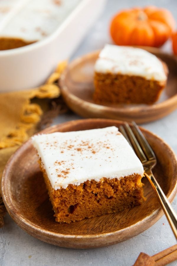 Two slices of pumpkin cake on small wooden plates with gold forks and the rest of the cake in the background.