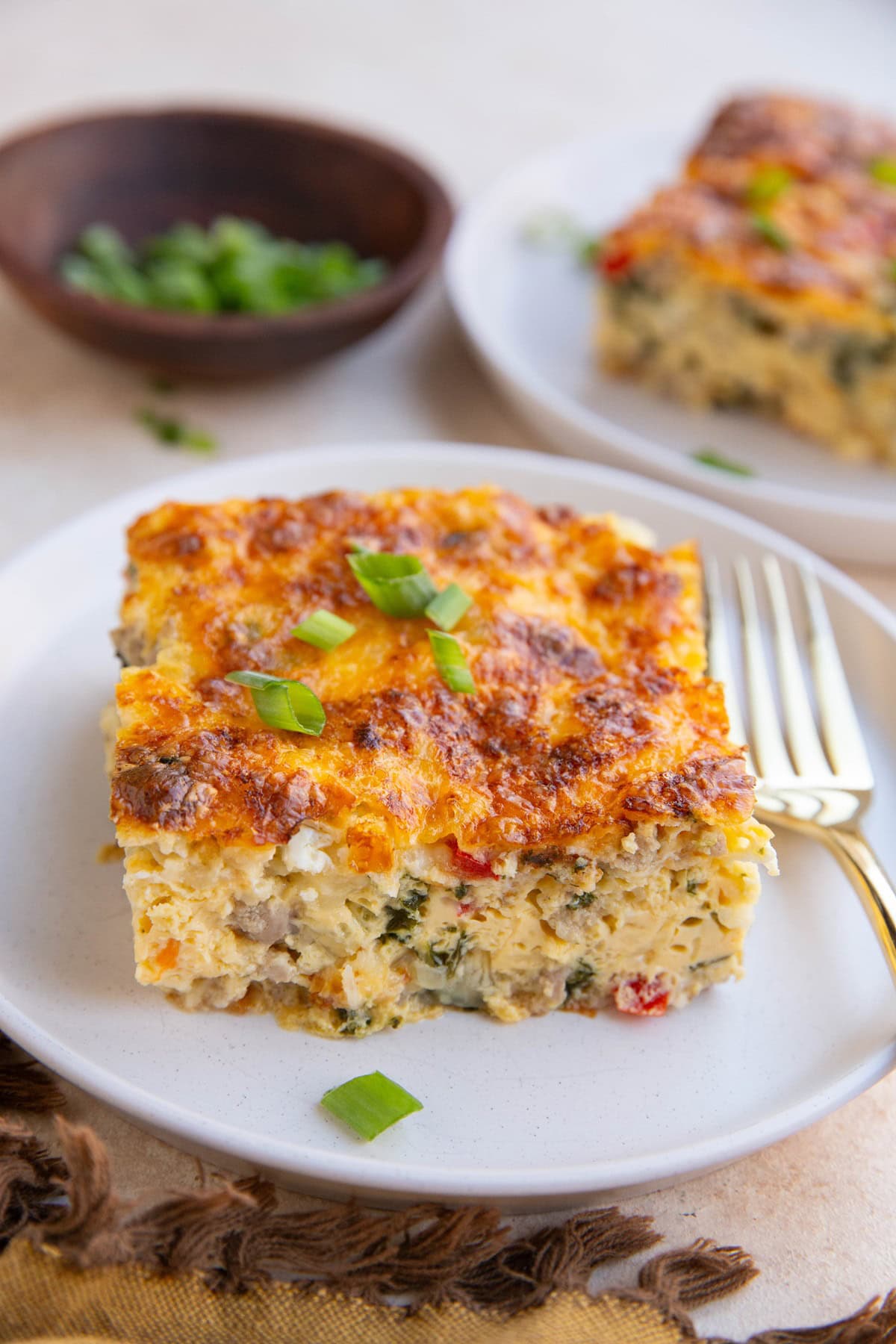 Two slices of breakfast casserole on two white plates with a bowl of chopped green onions in the background.