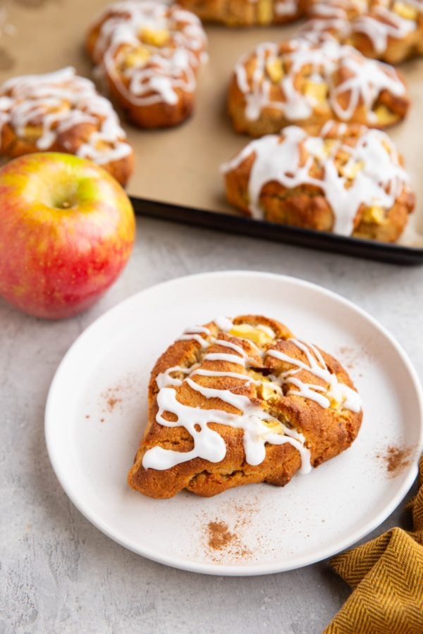 Plate with an apple scone on it with the baking sheet of scones in the background.