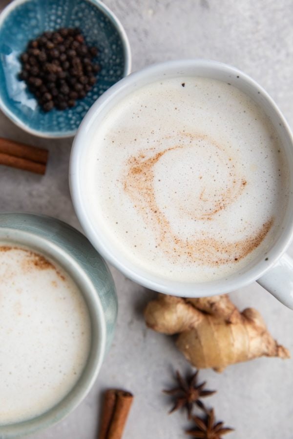 Two chai lattes on a table with spices all around, ready to drink.