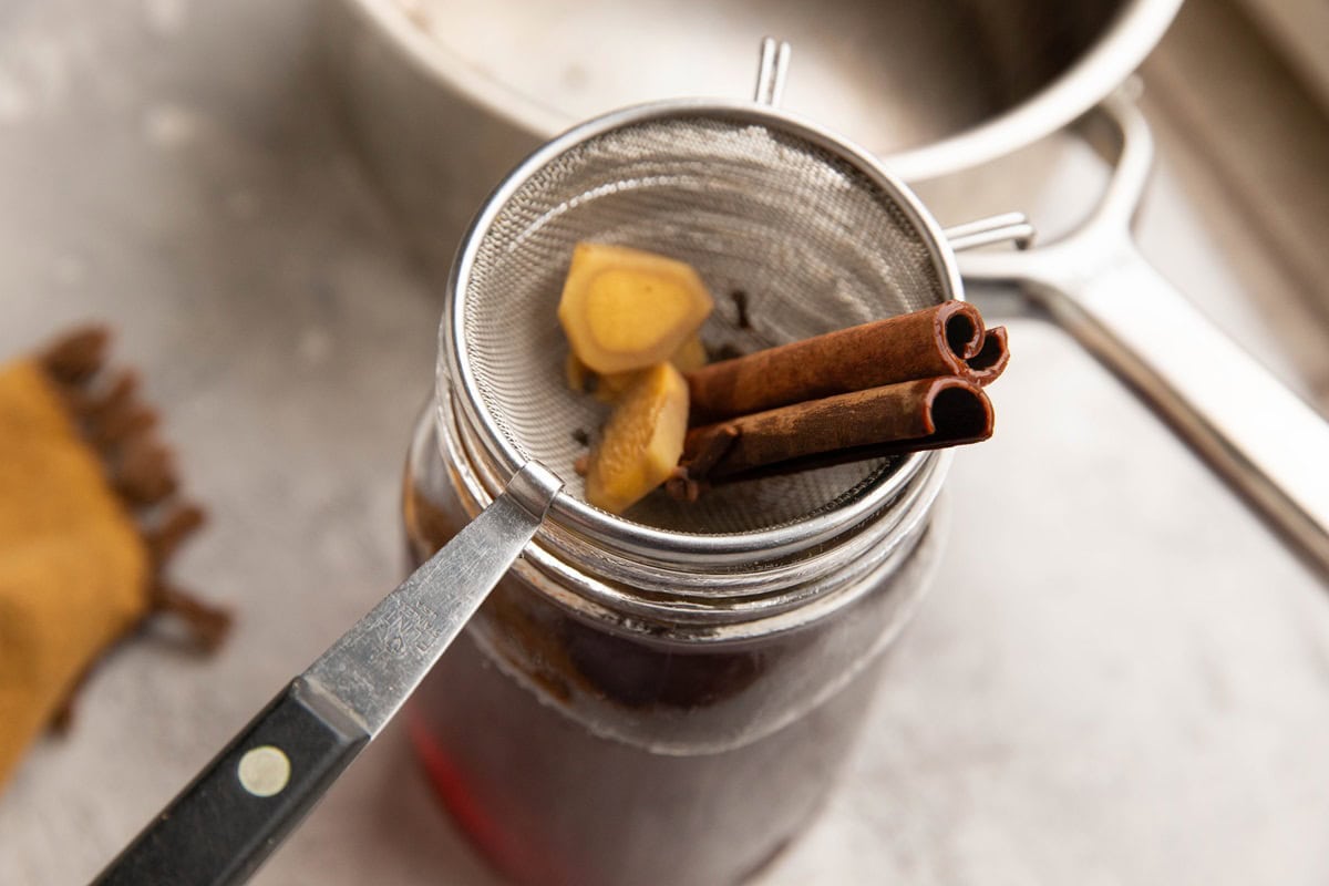 Small fine strainer over a large glass jar, straining the chai spices out of the tea