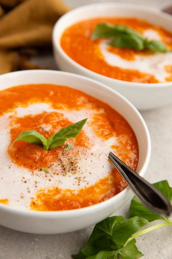 Two bowls of creamy tomato soup with a spoon and fresh basil leaves.