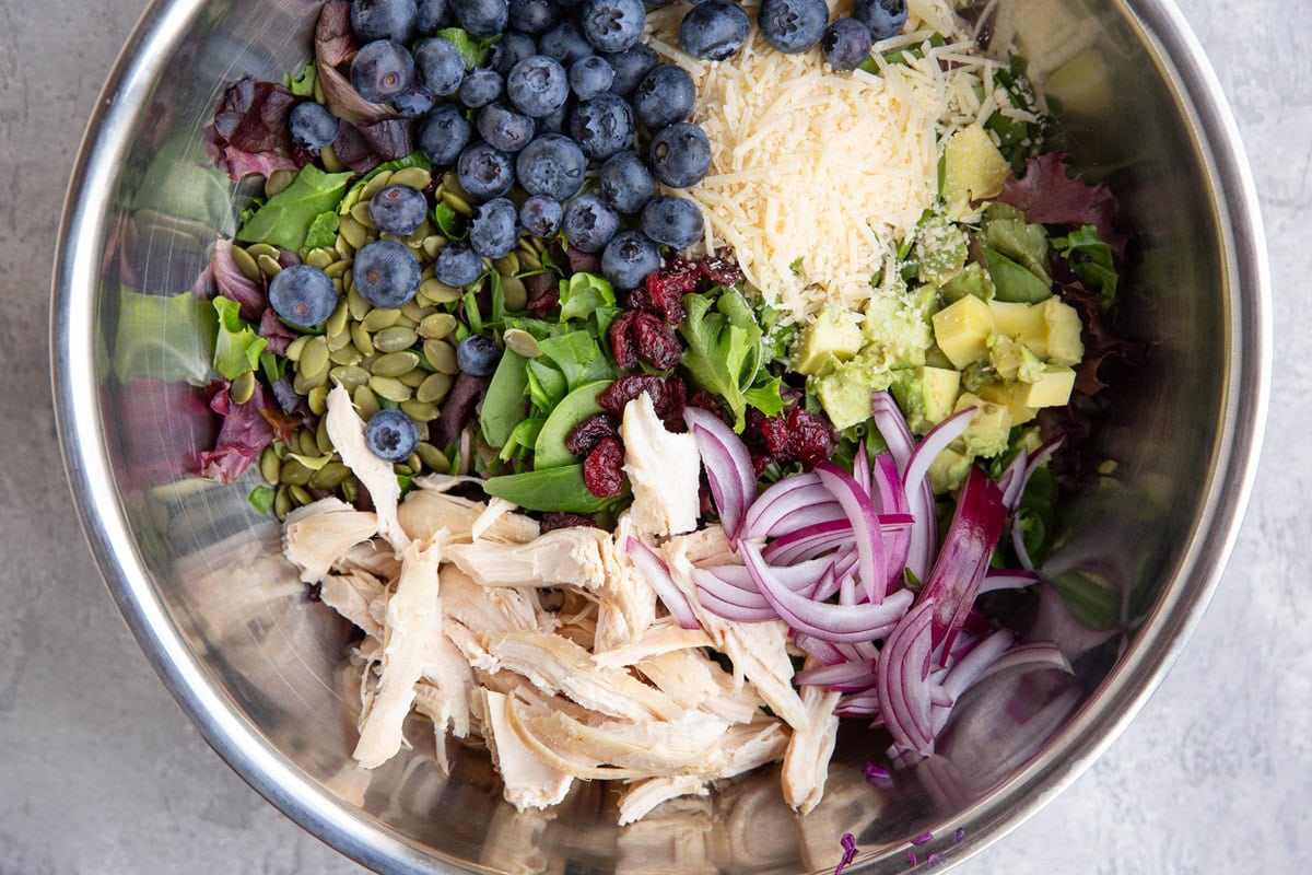 Large stainless steel bowl full of salad ingredients like chicken, blueberries, avocado, etc.