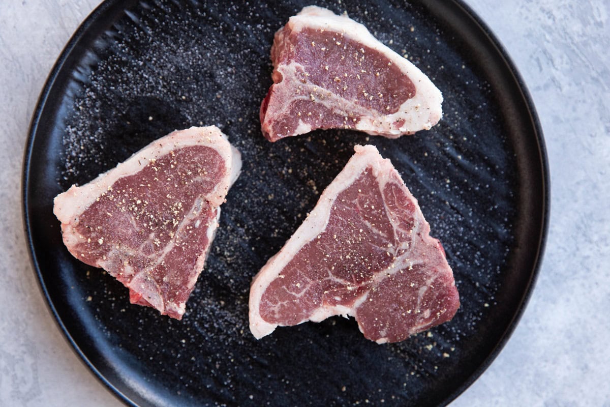 Black plate with raw pork chops on top, sprinkled with sea salt and black pepper.