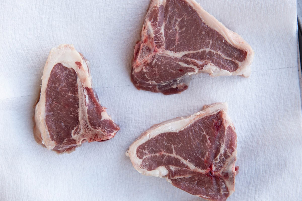 Plate with a paper towel on top and three raw lamb chops, patting the chops dry for cooking.