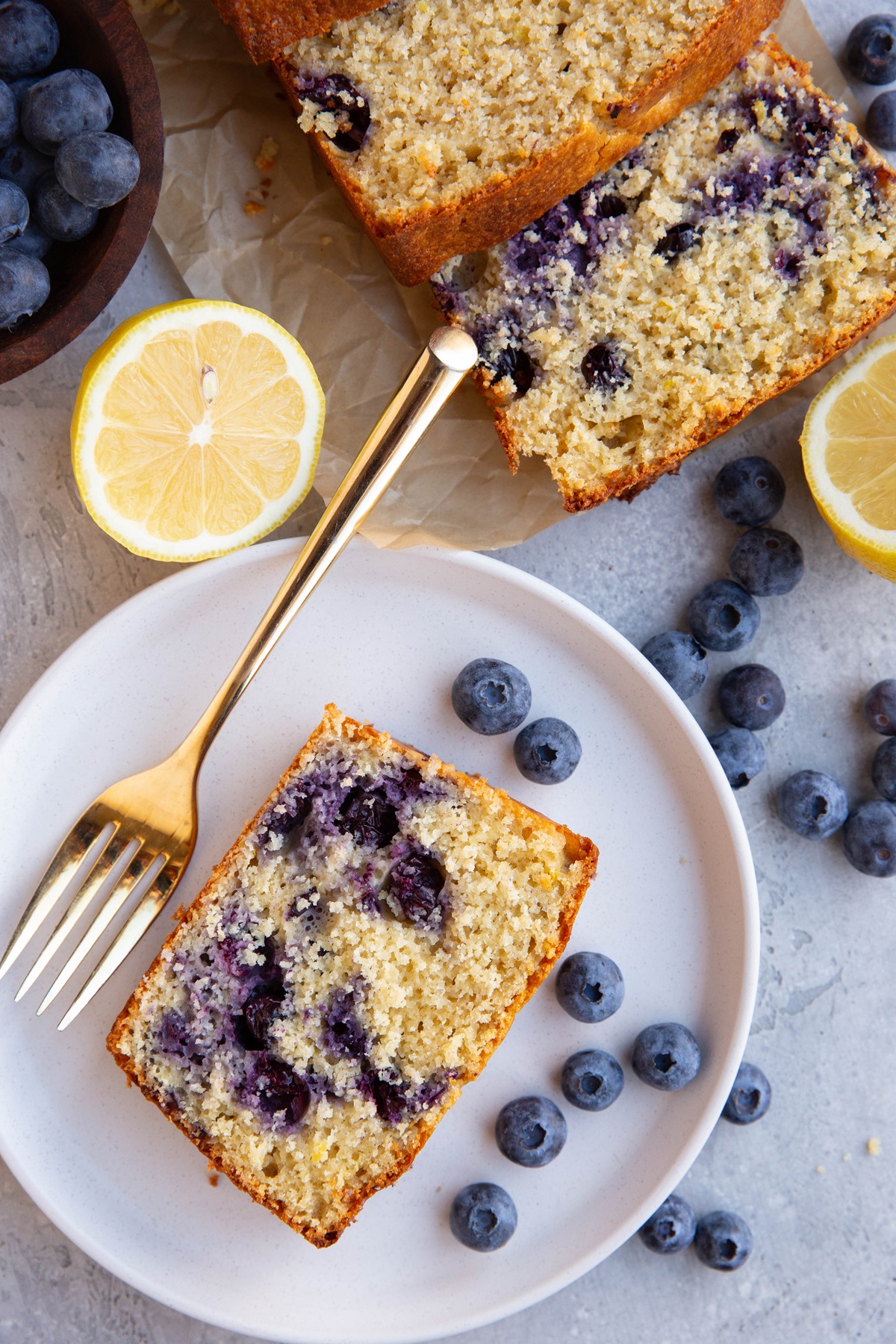 White plate with a slice of lemon blueberry bread and a loaf of blueberry bread to the side with fresh blueberries all around.